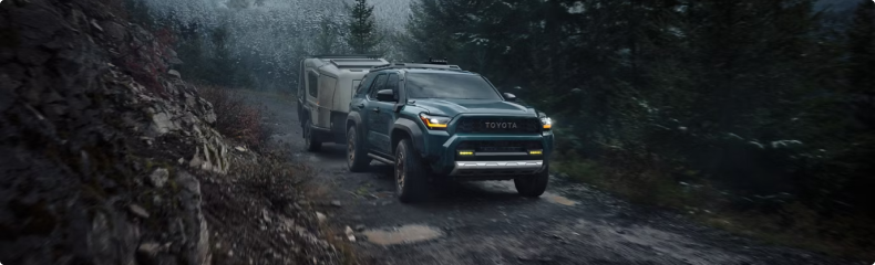 A green Toyota 4Runner towing a trailer up a wet gravel road through evergreen forest