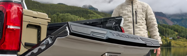 A man standing behind the open tailgate of a Toyota Tacoma beside a mountain lake