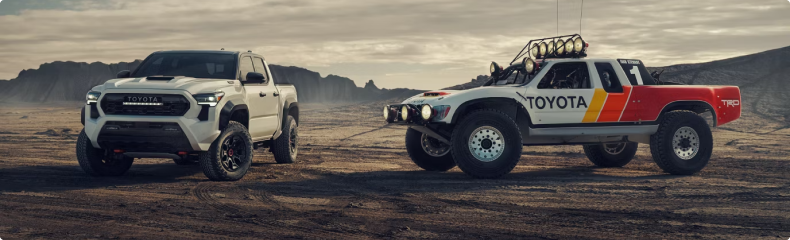 A white Toyota Tacoma beside a Toyota off-road race truck on a dusty plain