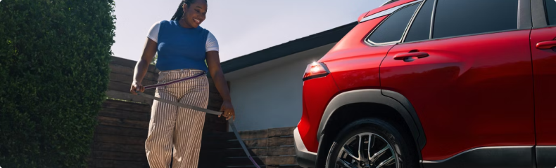 A woman coiling a garden hose next to a red Toyota Corolla Cross in a driveway