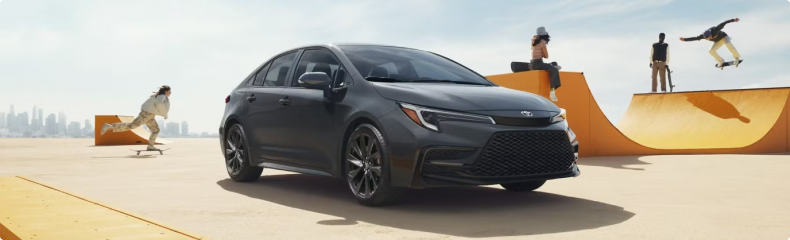 A gray Toyota Corolla parked among skateboard ramps with skaters riding behind it