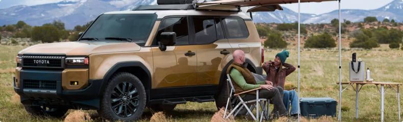 Two campers relaxing beside a tan Toyota Land Cruiser with a rooftop tent in a mountain meadow