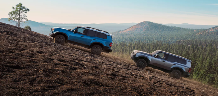 A blue and a silver Toyota Land Cruiser climbing a bare hillside above forested ridges