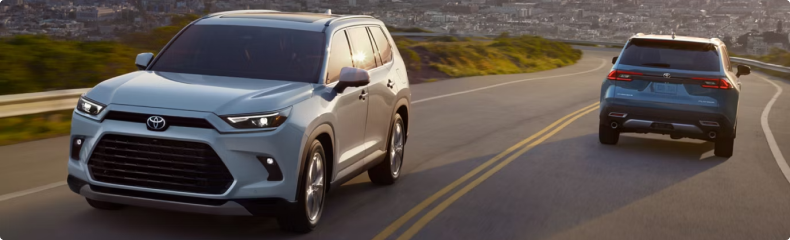 Two light blue Toyota Grand Highlanders passing each other on a winding road above a city