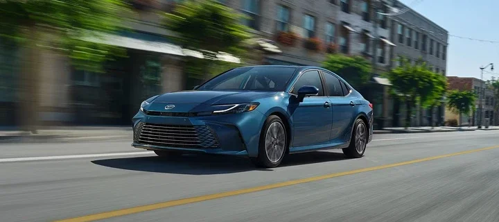 A blue Toyota Camry driving down a tree-lined town street