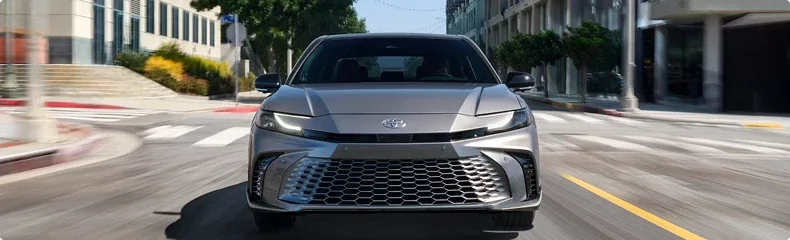 A silver Toyota Camry driving head-on down a sunlit city street