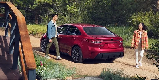 Two people walking toward a red Toyota Corolla parked on a dirt path beside a wooden footbridge