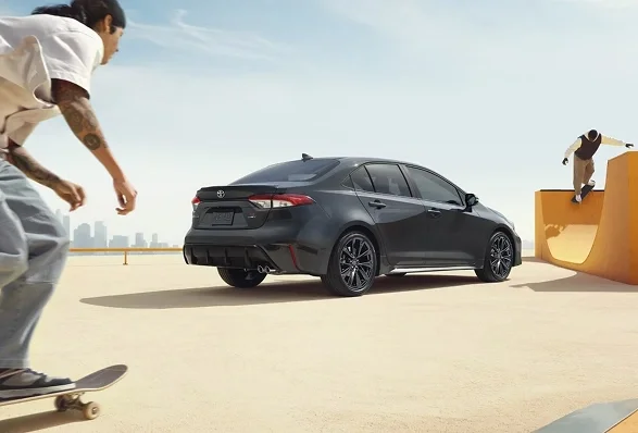 Dark gray Toyota Corolla parked at a sunny skate park while skateboarders ride nearby