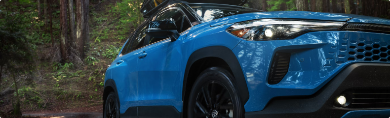 Close-up of the headlight and front fender of a blue Toyota Corolla Cross Hybrid in a forest