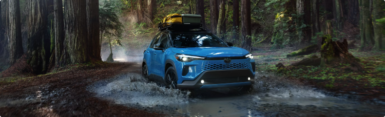 A blue Toyota Corolla Cross Hybrid with a roof cargo box splashing through a forest stream