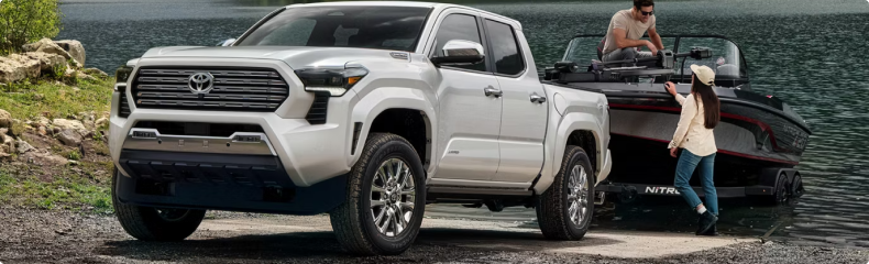 A white Toyota Tacoma at a lake boat launch with a boat and two people behind it