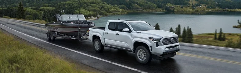 A white Toyota Tacoma towing a boat on a wet highway beside a lake