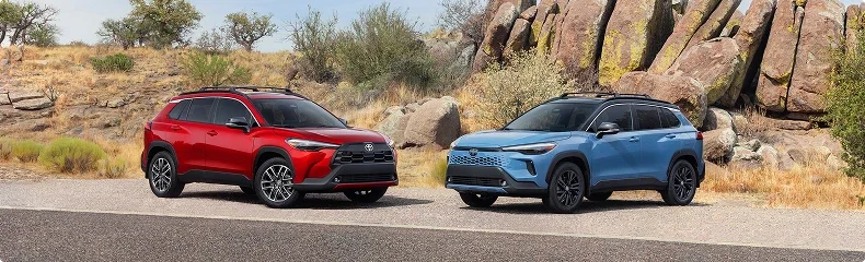 A red and a blue Toyota Corolla Cross parked side by side on a roadside beside rock outcrops