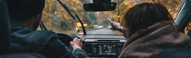 View from the back seat of a driver and passenger on a rain-streaked two lane road through autumn woods