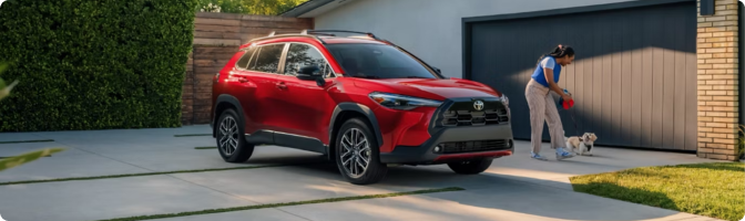 Red 2026 Toyota Corolla Cross parked in a driveway while a woman plays with a small dog by the garage