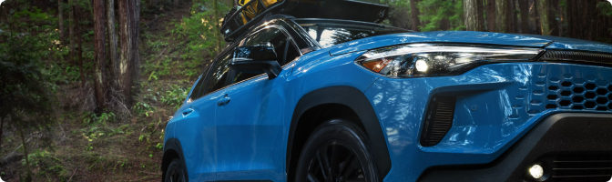 Blue 2026 Toyota Corolla Cross Hybrid with a roof cargo box parked in a forest