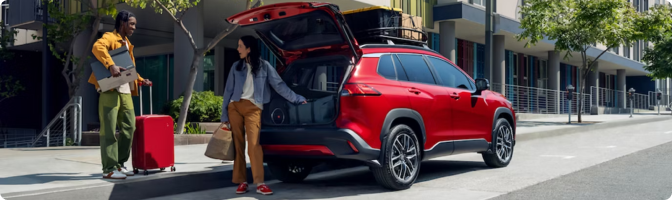 Two people loading luggage into the open liftgate of a red 2026 Toyota Corolla Cross on a city street