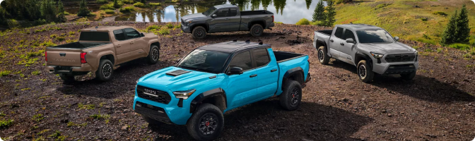 Four 2026 Toyota Tacoma trucks in different colors parked together on a trail near a lake