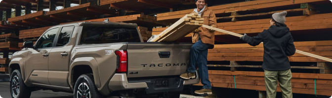 Two people loading lumber into the bed of a tan 2026 Toyota Tacoma at a lumber yard