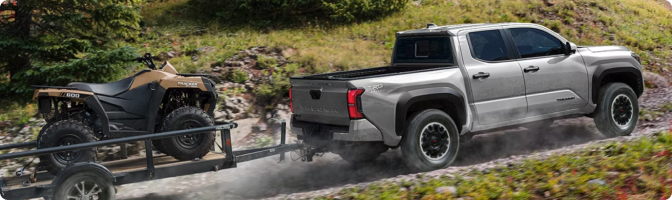 Silver 2026 Toyota Tacoma towing an ATV on a utility trailer along a gravel road